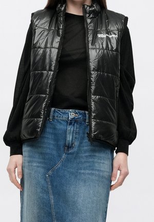 Personne portant un gilet matelassé noir brillant avec le logo Karl Lagerfeld Jeans, une chemise noire à manches longues et une jupe en jean bleue, debout devant un fond blanc.