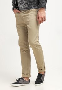 Homme portant un pantalon beige slim-fit avec des ourlets retroussés, des chaussures à lacets noires et grises, une chemise noire à motifs et une veste en cuir noire.