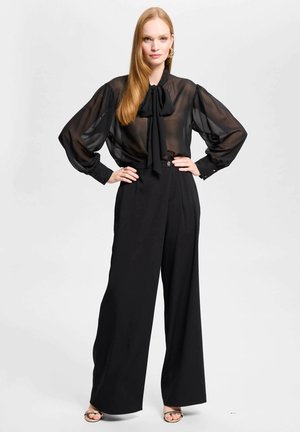 Vrouw staat met handen in de zij, draagt een doorschijnende zwarte blouse met strik en hoog getailleerde wijde zwarte broek tegen een effen achtergrond.