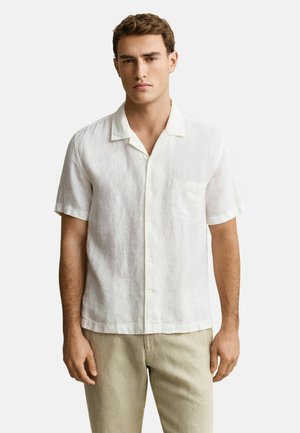 Homme portant une chemise blanche à manches courtes avec une poche poitrine et un pantalon beige, debout devant un fond clair uni.