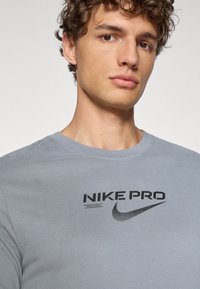 Lys grå crewneck t-skjorte laget av mykt stoff. Har en svart "NIKE PRO"-logo og swoosh trykt på brystet. Glatt tekstur.