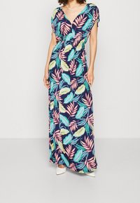 Kobieta w granatowej maxi sukience z różowymi, żółtymi i zielonymi wzorami tropikalnych liści oraz białych czółenkach na jednolitym tle.