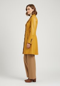 Yellow leather coat with a tailored fit, featuring buttoned cuffs and a single chest pocket, paired with beige wide-leg trousers and brown heels.