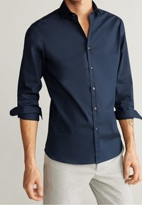 Camicia a maniche lunghe blu navy con colletto abbottonato, caratterizzata da una texture liscia e polsini arrotolabili, abbinata a pantaloni grigio chiaro.