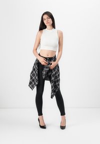 Witte mouwloze crop top, zwarte high-waisted jeans, zwart-grijze geruite blouse vastgebonden om de taille en zwarte hoge hakken voor een effen achtergrond.