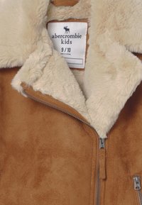 Giubbotto in suede marrone con colletto e fodera in pelliccia sintetica beige morbida. Include una cerniera frontale e un'etichetta con le informazioni sulla taglia.