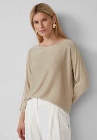 Beige, strukturiertes Langarmoberteil mit Bootshals, kombiniert mit weißen, plissierten Hosen. Verfügt über einen dezenten Glanz und einen lässigen Schnitt.