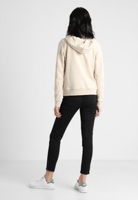 Donna con capelli scuri che indossa una felpa leggera beige, pantaloni aderenti neri con cerniere alla caviglia e scarpe da ginnastica bianche, in piedi di spalle su uno sfondo bianco.