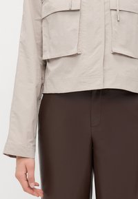 Light beige cropped jacket with two front pockets, features a smooth texture and drawstring closure. Paired with brown pants.
