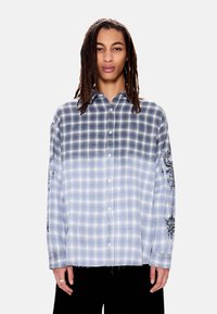 Νεαρός με μακριές πλεξούδες ντραντλoκ που φοράει μπλε και λευκό πουκάμισο με σχέδιο καρό σε διαβάθμιση, με μαύρα γραφικά στο μανίκι.