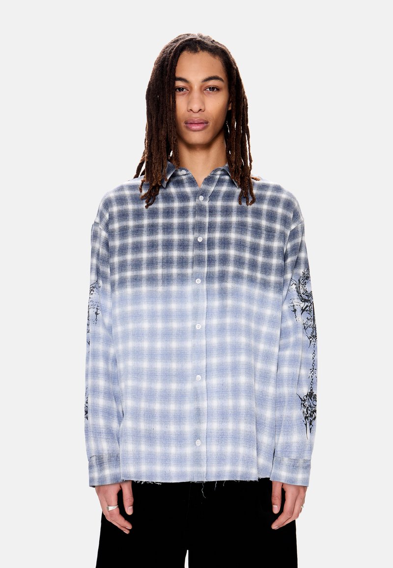 Νεαρός με μακριές πλεξούδες ντραντλoκ που φοράει μπλε και λευκό πουκάμισο με σχέδιο καρό σε διαβάθμιση, με μαύρα γραφικά στο μανίκι.