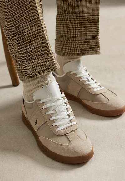 Beige en witte veter sneakers met bruine zolen, gedragen met crèmekleurige sokken en bruin geruite broek, voeten rustend op een beige vloer.