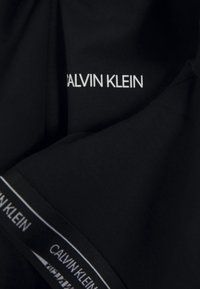 Zwarte stof met het "CALVIN KLEIN" logo in het wit, gepositioneerd op de borst. De tailleband heeft een herhalend logo-patroon, wat de zichtbaarheid van het merk vergroot.