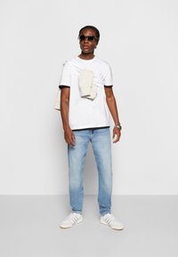 Maglietta bianca con dettagli neri, jeans azzurri chiari, maglione beige drappeggiato sulle spalle e occhiali da sole neri con trama. Sneakers bianche con strisce.