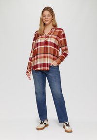 Rood en beige geruit overhemd met lange mouwen, knoopsluiting aan de voorkant en een relaxte pasvorm, gecombineerd met blauwe spijkerbroeken en beige sneakers.