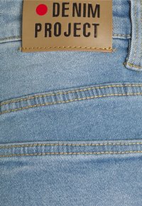 Světle modrá dením tkanina s viditelnou texturou, opatřená hnědou koženou etiketou s nápisem "DENIM PROJECT" v tučném černém písmu s červeným akcentem.