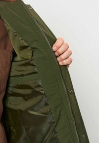 Main tenant ouverte une veste vert olive non zippée montrant sa doublure intérieure lisse et brillante ainsi que les détails de la fermeture éclair sur un fond uni.