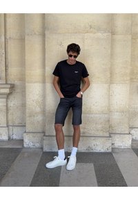 Schwarzes T-Shirt, dunkle Jeansshorts, weiße Sneakers mit weißen Socken, steht an einer strukturierten Steinmauer. Lässiges Urban-Outfit, lockere Passform.