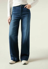 Szerokie, ciemnoniebieskie dżinsy z denimu, z wysokim stanem, przednimi kieszeniami i delikatnym wyblaknięciem. Połączone z jasnymi sneakerami.