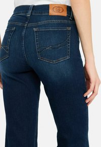 Jeans de mezclilla azul oscuro con cintura ajustada, parche de cuero en la parte trasera y costuras decorativas con rhinestones en los bolsillos.
