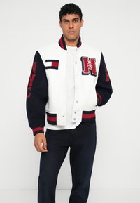 Balta varsity striukė su tamsiai mėlynomis rankovėmis, raudonais akcentais ir siuvinėtomis raidėmis. Dėvima ant baltos marškinėlių, derinama su tamsiomis džinsomis.