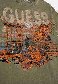 Olīvu zaļš kokvilnas džemperis ar lieliem burtiem "GUESS". Iezīmēts ar spilgti oranžu izšuvumu ar ziediem un abstraktiem dizainiem.