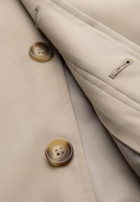 Tessuto di un trench coat beige con una superficie liscia, dotato di un grande bottone marrone e un dettaglio sul colletto cucito.