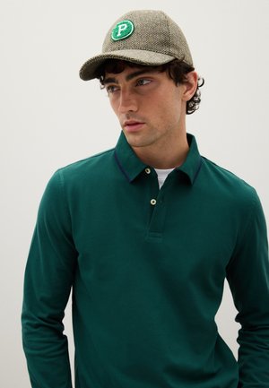 Groen poloshirt met lange mouwen en een kraag met een paarse accent. Het model draagt een bruine visgraatpet met een groen logo.
