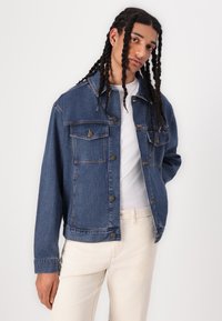 Jeans jakna v srednji modri barvi, z gumbnimi zapiranji, žepoma na prsih in sproščenim krojem. Nosi se čez belo srajco z svetlimi hlačami.