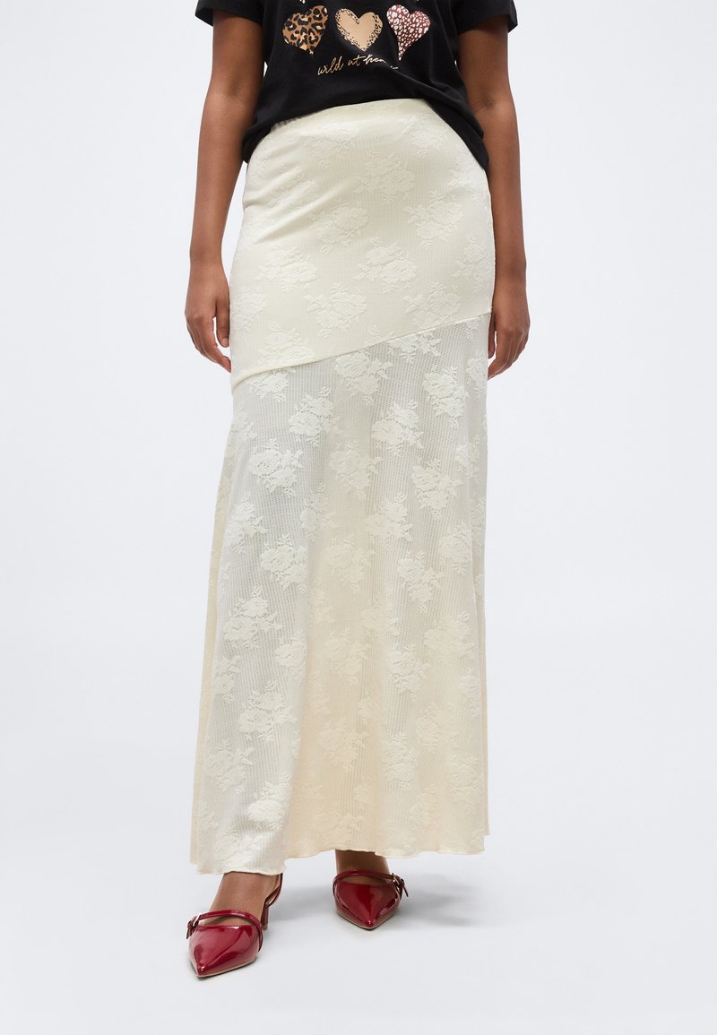 Γυναίκα που φοράει μια εκρού maxi φούστα από δαντέλα με λουλούδια και ασύμμετρο τελείωμα και κόκκινα μυτερά τακούνια, σετ με ένα μαύρο μπλουζάκι με γραφικό σχέδιο.