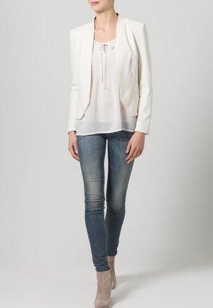 Blazer bianco con un design aderente, abbinato a una blusa bianca trasparente e jeans skinny blu, completato da stivaletti beige alla caviglia.