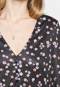 Blouse noire à motif floral avec un décolleté en V, ornée de petites fleurs blanches, violettes et orange sur un fond à pois, avec des boutons noirs.