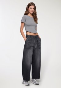 T-shirt grigia corta e jeans neri a gamba larga con tasche frontali, caratterizzati da un design sbiadito. Sneakers argentate completano l'outfit.