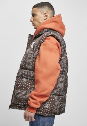 Bočný pohľad na osobu, ktorá má na sebe hnedú vzorovanú páperovú vestu cez oranžovú mikinu s kapucňou a modré džínsy na jednofarebnom pozadí.