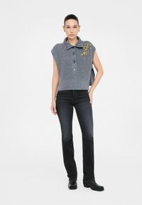 Gilet grigio in maglia con colletto, caratterizzato da tre bottoni e decorazioni floreali gialle. Abbinato a jeans scuri e stivaletti neri.