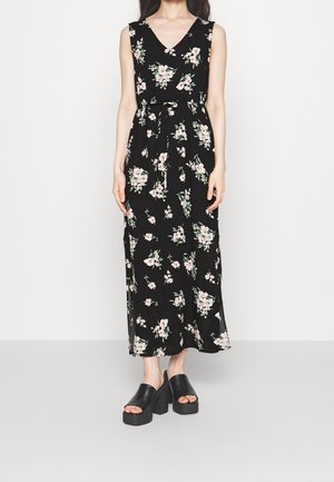 Femme portant une robe maxi noire sans manches avec un imprimé floral rose et blanc et une ceinture à la taille, associée à des sandales compensées noires.