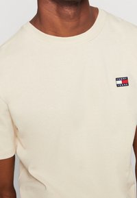 T-shirt in cotone beige con collo rotondo e maniche corte. Include una piccola toppa con logo ricamato in rosso, bianco e blu.