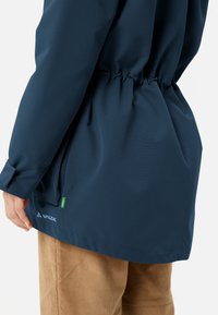 Blauwe waterdichte jas met een taille die kan worden aangesnoerd, lange mouwen en een gladde textuur. Bevat een subtiele logo en een groene accentlabel aan de onderkant.