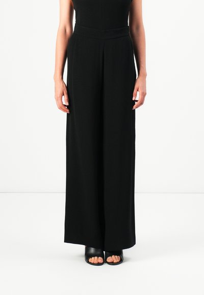 Donna che indossa pantaloni neri a vita alta e dal taglio ampio con una cintura aderente e scarpe con tacco aperte nere su uno sfondo bianco.