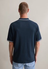 Granatowa koszulka polo z teksturowanej tkaniny, krótkie rękawy oraz białe paski na kołnierzyku i mankietach, widoczna z tyłu.