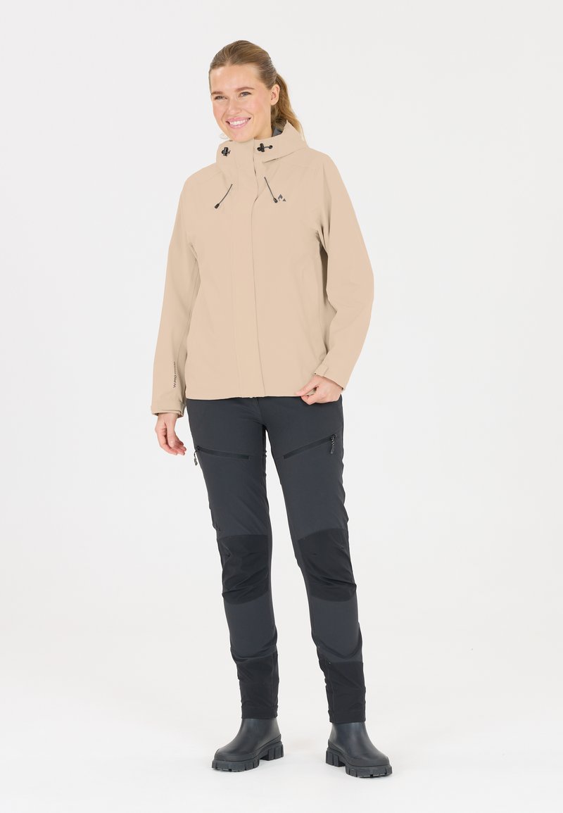 Ljusbeige vattentät jacka med justerbar huva, tillsammans med svarta byxor med blixtlåsfickor och kontrasterande paneler. Svarta stövlar ingår.