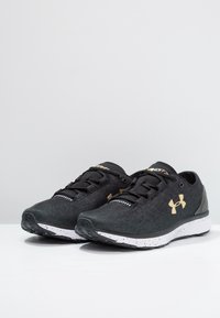 Chaussures de sport noires avec une tige texturée, des accents dorés, une semelle blanche mouchetée, et des lacets noirs. Design léger et épuré.