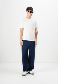T-shirt en coton blanc, jean en denim foncé ample, et lunettes de soleil noires. Baskets avec des accents blancs et gris. Fond uni.