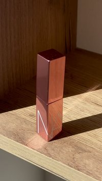 Rektangulär läppstiftsbehållare med en reflekterande rosaguldfinish, som har en minimalistisk vit geometrisk design. Placerad på en träytan.