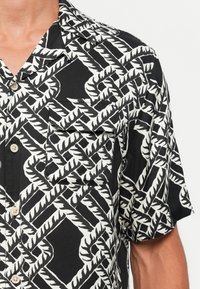 Black and white short-sleeve shirt with a bold diagonal leaf pattern and front button closure on a person’s upper torso.