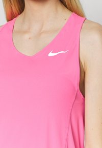 Růžový bezrukávový sportovní top s výstřihem do V, vyrobený z hladké látky. Na levé části hrudi je bílá ikona Nike.