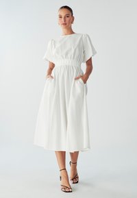 Biała lniana sukienka z elastycznym pasem, krótkimi bufiastymi rękawami, kieszeniami z boku oraz zwiewnym krojem, w zestawie z czarnymi sandałami na obcasie.