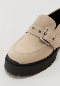 Beige Wildleder Slipper mit einem Rutschdesign, ausgestattet mit einem breiten Riemen, einer silberfarbenen Schnalle und einer dicken schwarzen Plateausohle.