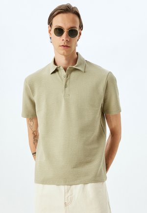 Camisa polo de textura de gofre en verde claro, con cuello clásico, tres botones y mangas cortas. Se lleva con gafas de sol redondas.