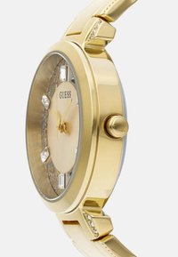 Zlatý náramkový hodinky s okrúhlym puzdrom, hodinovými ukazovateľmi zdobenými kryštálmi a hladkým rámčekom. Remienok je lesklý v zodpovedajúcej zlatej farbe.
